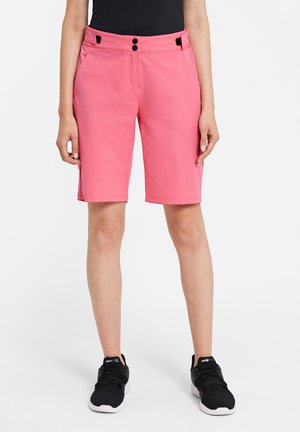 Woman wearing pink knee-length shorts with black buttons, black sleeveless top, and black athletic shoes standing against white background.