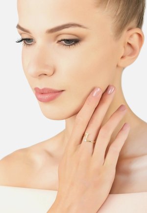 Gold ring with a 'T' design, featuring a small decorative stone, worn on a hand touching the cheek. Smooth skin, soft nail polish.