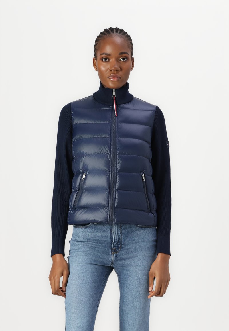 Gilet imbottito blu navy con colletto alto, zip frontale e tasche laterali, caratterizzato da un design trapuntato e una finitura lucida e raffinata. Abbinato a un top scuro.