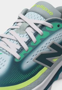 Κοντινό πλάνο αθλητικού παπουτσιού σε ανοιχτό μπλε και πράσινο χρώμα με άσπρα κορδόνια, μαξιλαρωμένη γλώσσα με ετικέτα "ellipse" και μεγάλο λογότυπο "N" στο πλάι.