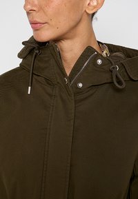Olijfgroene jas met hoge kraag, rits en drukknoopsluiting, en een trekkoordcapuchon. Kenmerkt zich door een gladde, gestructureerde stof.