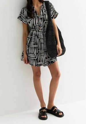 Žena v krátkých šatech s černo-bílým vzorem, v černých sandálech, s velkou černou taškou z háčkované příze přes rameno.