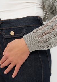 Χέρι που ακουμπά στην τσέπη σκούρου μπλε τζιν, φορώντας μπεζ πλεκτό μακρυμάνικο τοπ με δαντελένιο μοτίβο και ριμπ μανσέτα.