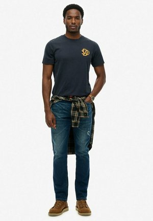 Marine T-shirt met gouden logo, blauwe jeans en bruine laarzen. Geruite shirt om de taille geknoopt; denim jeans hebben lichte slijtage details.