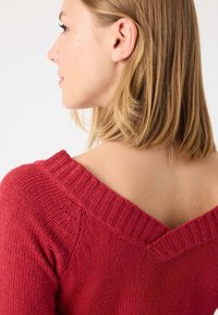 Suéter de punto rojo con diseño de cuello en forma de V, cuello acanalado y tejido texturizado. Parte de la espalda del modelo es visible, con cabello castaño claro y largo hasta los hombros.