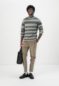 Pull à col roulé gris rayé, pantalon beige plissé, sac à main noir, chaussures noires, debout contre un fond blanc.