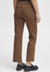 Pantalon marron, extensible, avec une coupe slim, doté d'une ceinture, de deux poches arrière et de jambes droites. Porté avec des chaussures noires imposantes.