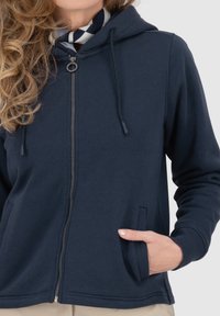 Felpa con zip di colore navy realizzata in tessuto morbido. Presenta un cappuccio regolabile con cordino, tasche laterali e polsini a costine. Superficie liscia con dettagli minimali.