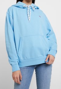 Jasnoniebieska bluza z kapturem wykonana z miękkiego materiału, z przednią kieszenią, ściągaczami na mankietach i wzorzystym ściągaczem w kapturze.