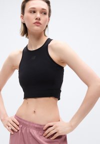 Νεαρός με κοντά μαλλιά που φοράει μαύρο αμάνικο crop top και μωβ αθλητικές σορτς, στέκεται με τα χέρια στη μέση, κοιτώντας πλάγια.