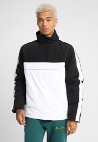 Half-zip pullover laget av et slitesterkt stoff, med en svart toppseksjon, hvit kropp og sidestriper, samt høy krage.