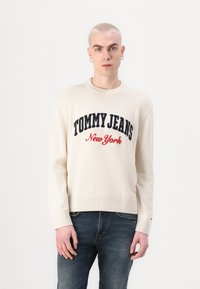 Pulover crem cu tivuri și manșete cu nervuri, av�ând broderie în albastru închis și roșu "TOMMY JEANS New York", asortat cu blugi de culoare albastru închis.