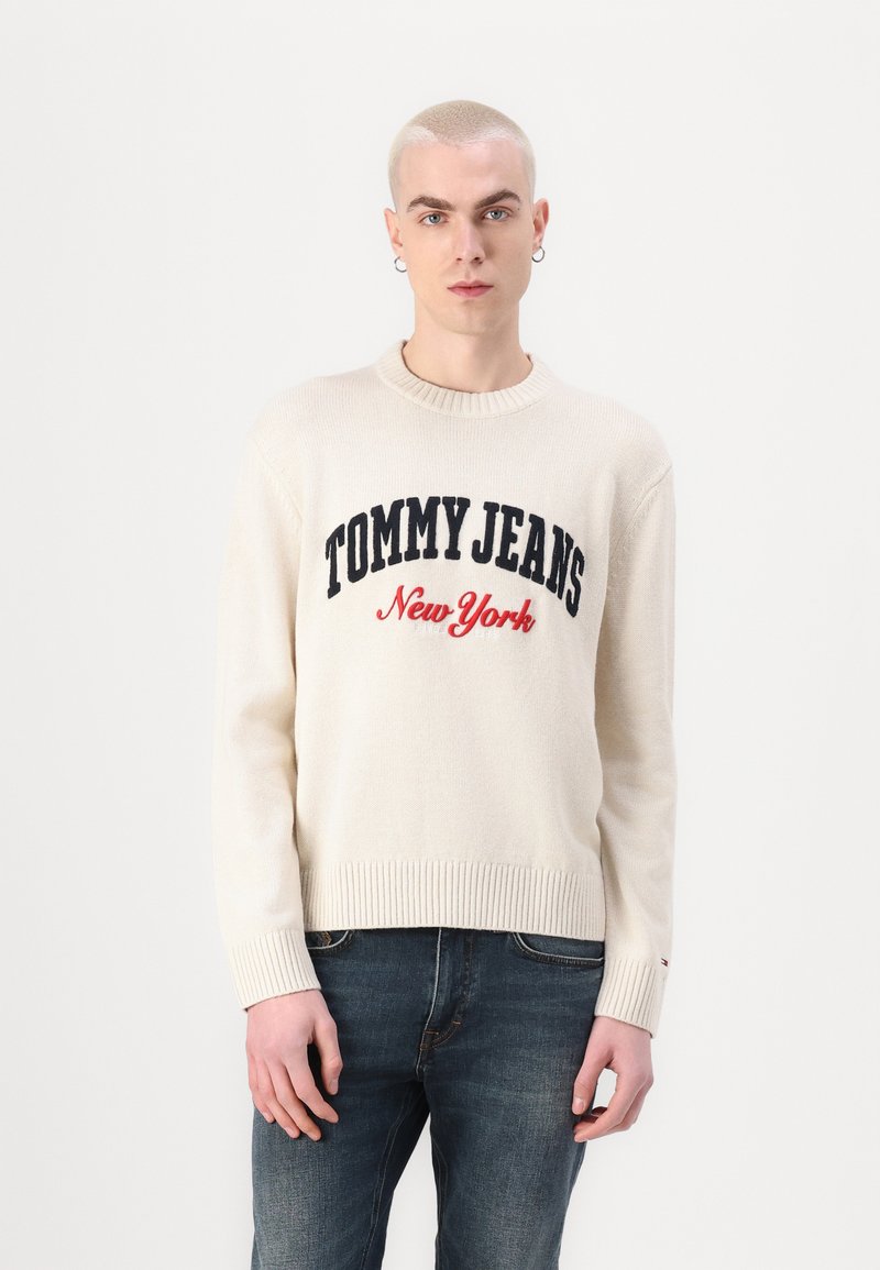 Kremgenser med ribbestrikket kant og mansjetter, med "TOMMY JEANS New York" i marineblå og rød brodering, kombinert med mørkeblå jeans.