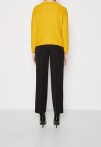 Gul stickad tröja med en kortad design, bärs över svarta högmidjade byxor och svarta klackade ankelstövlar, sedd bakifrån.