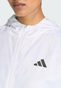 White lightweight jacket with a zip front, adjustable hood, and a black triangular logo on the left chest. Smooth texture, minimalist design.