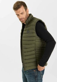 Olivgrön vadderad väst med vertikal quiltad textur, ärmlös design och dragkedjestängning. Kombinerad med en svart långärmad tröja och denimjeans.