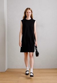 Femme portant une robe noire sans manches, des chaussettes blanches, des chaussures Mary Jane noires, portant un sac à main noir, marchant sur un sol en bois contre un mur uni.