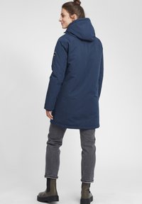 Mørkeblå isolert jakke med hette, med rett snitt, frontglidelås og emblemdetalj på ermet. Brukt med grå jeans og grønne støvler.