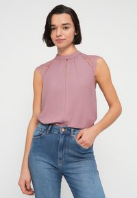 Αμάνικη μπλούζα σε αποχρώσεις σάπιο ρόδο με δαντελένιες λεπτομέρειες στους ώμους, λαιμόκοψη με άνοιγμα και μαλακό ύφασμα, σε συνδυασμό με ψηλόμεσα μπλε τζιν.