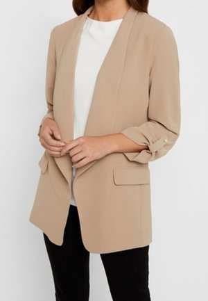 Kobieta ubrana w beżową marynarkę z otwartym przodem i podwiniętymi rękawami, na białym topie i czarnych spodniach na jednolitym tle.