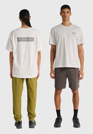 Two models wear white graphic t-shirts; one faces forward with gray shorts, the other faces backward with olive green joggers and black sneakers.