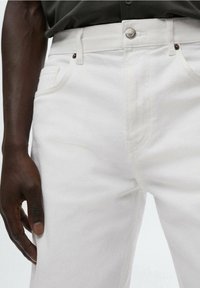 Persona de piel oscura vistiendo jeans de mezclilla blancos y una camiseta oscura, mostrando una mano descansando junto al muslo y los detalles frontales de los jeans.