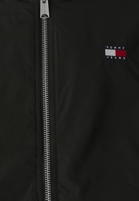 Svart vindjakke med sølvglidelås. Har en liten brodert logo i marineblått, rødt og hvitt på brystet. Glatt tekstur.
