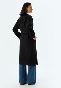 Zwarte lange jas met een strakke taille, brede mouwen en zijzakken, gecombineerd met blauwe wijde jeans en hoge hakken.