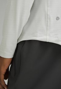 Helle graue Langarmbluse aus strukturiertem Stoff mit detaillierter Naht, kombiniert mit schwarzen Hosen mit einer Seitentasche. Logo sichtbar am Saum des Shirts.