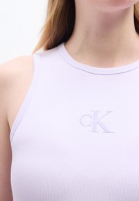 Κοντινό πλάνο ενός ατόμου που φοράει μωβ αμάνικη μπλούζα με ριγέ υ φή και διακριτικό λογότυπο Calvin Klein στο στήθος.