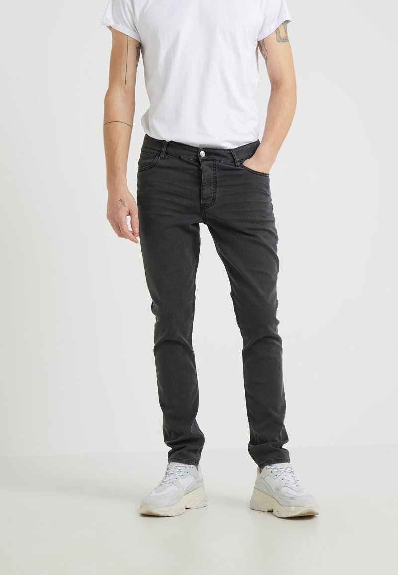 Homme portant un jean noir slim, un t-shirt blanc et des baskets blanches, debout avec une main dans la poche devant un fond uni.