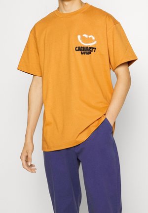 Homme portant un t-shirt orange avec un smiley blanc et le texte noir « CARHARTT WIP », associé à un pantalon bleu, main dans la poche.