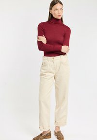 Burgunderfarbener Rollkragenpullover kombiniert mit cremefarbenen, hochgekrempelten Hosen. Das Model trägt Loafers mit Leopardenmuster. Einfacher, entspannter Schnitt.