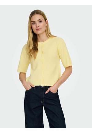 Frau trägt eine gelbe kurzärmelige, geknöpfte Strickjacke und dunkelblaue, hoch taillierte Jeans, steht mit den Händen in den Taschen.