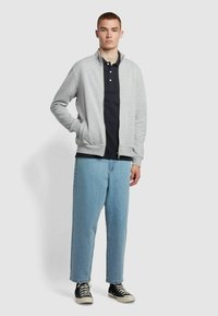 Szara kurtka zapinana na zamek z wysokim kołnierzem, noszona na granatowej koszulce polo. Jasnoniebieskie dżinsy o luźnym kroju oraz czarne sneakersy dopełniają strój.