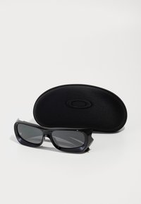 Óculos de sol retangulares pretos com lentes escuras, posicionados em frente a uma caixa rígida oval preta, sobre um fundo branco.