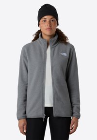 Persona con capelli ricci che indossa un berretto nero, una giacca in pile grigia con zip, una maglietta bianca sotto e pantaloni neri, in piedi su uno sfondo semplice.