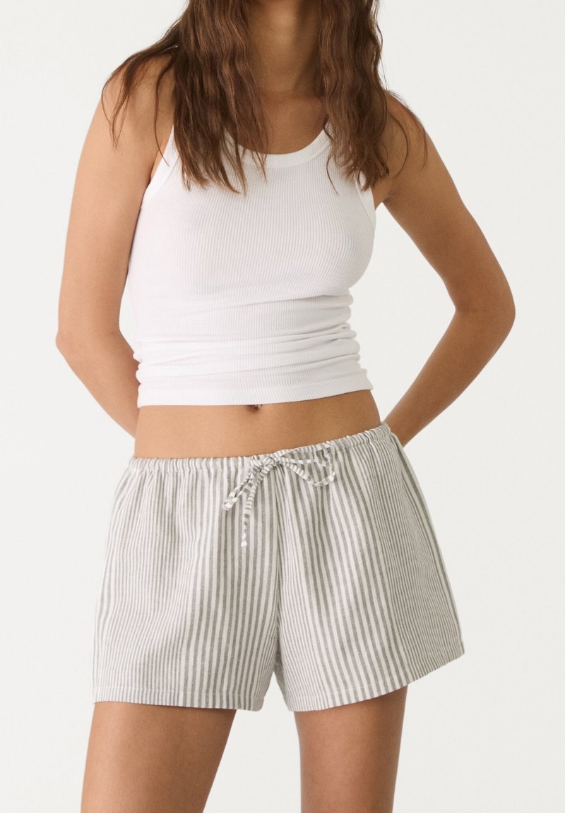 Femme portant un débardeur blanc côtelé et un short à cordon rayé gris clair et blanc, debout les mains derrière le dos.