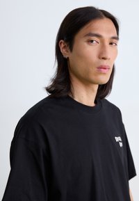 T-shirt en coton noir à manches courtes, col rond, avec un petit logo blanc imprimé sur la poitrine gauche. Texture lisse, coupe décontractée.