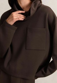 Brązowy sweter z kapturem i kieszenią na piersi, wykonany z miękkiego materiału. Posiada długie rękawy i luźny krój, eksponując gładką teksturę.