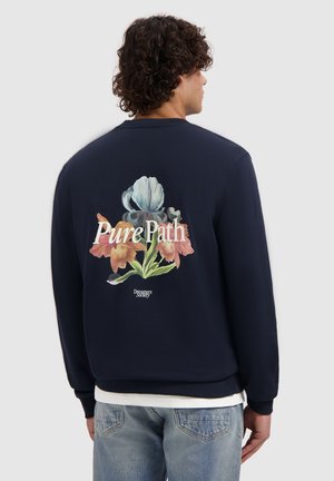 Man with curly hair wearing a navy sweatshirt featuring a large floral design and text "Pure Path" on the back, paired with light blue jeans.