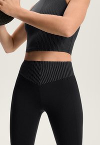 Crni sportski kratki top s bijelim dijagonalnim prugama u kombinaciji s visokim crnim tajicama koje imaju sličan uzorak pruga na struku.