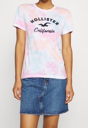 Kobieta ubrana w pastelowy t-shirt tie-dye marki Hollister California w połączeniu z niebieską jeansową spódnicą, stojąca na jednolitym tle.