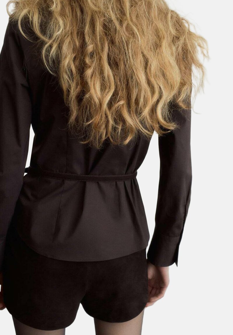 Zwarte blouse met een getailleerde taille, lange mouwen en gladde stof, gecombineerd met bruine suède shorts. Het haar is lang en golvend.