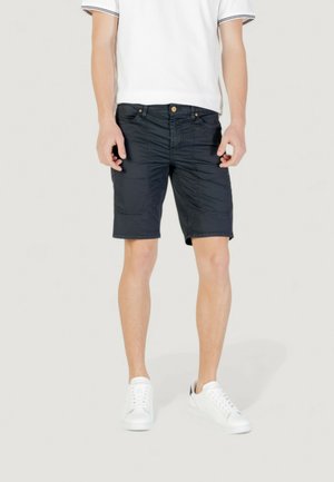 Pantaloni corti blu navy con una texture liscia, dotati di chiusura con zip e tasche frontali. Indossati con una camicia bianca e sneakers bianche.