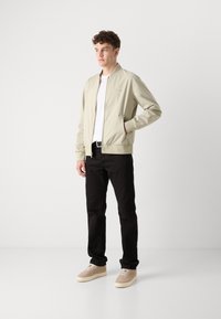 Hellbeige Bomberjacke mit einem Frontreißverschluss, kombiniert mit schwarzen Jeans und hellbeigen Sneakern, stehend vor einem weißen Hintergrund.