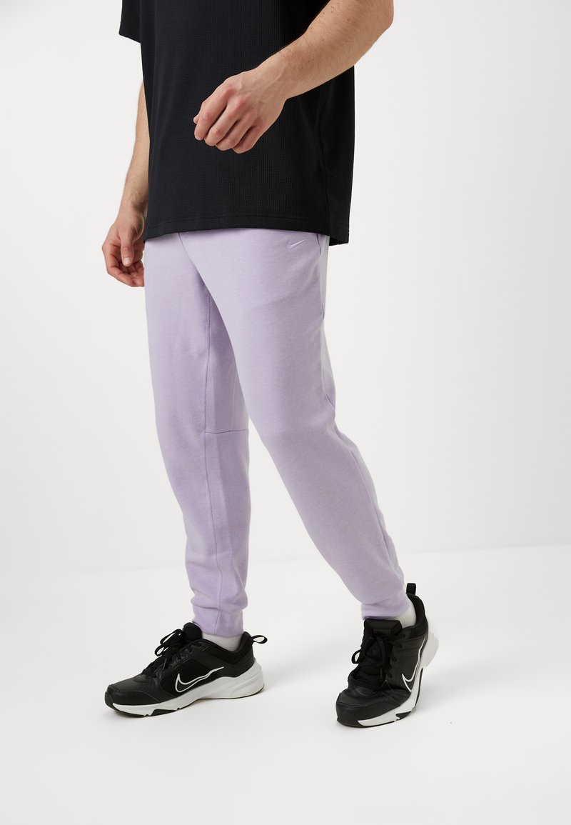 Lavendelfarbene Sporthose im Slim-Fit-Schnitt, aus einem Baumwoll-Mischgewebe gefertigt, kombiniert mit einem schwarzen strukturierten T-Shirt und schwarzen Sneakers.