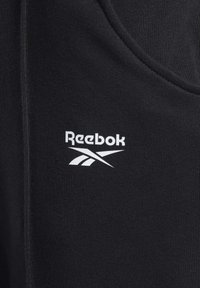 La tela negra presenta un acabado texturizado con un logo de Reebok en blanco y acentos de diseño. La prenda incluye una costura a lo largo del borde.