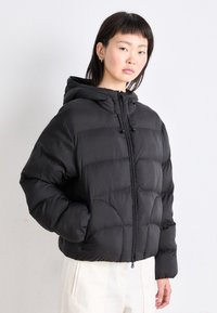 Melns pūkains jaka ar kapuci, ar quilted dizainu, priekšējā rāvējslēdzēja aizdare un elastīgi aproces. Mīksta, gluda tekstūra.