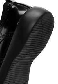 Zwarte sneaker met een glanzend leren bovenwerk, een getextureerde rubberen zool en gestikte details aan de zijkanten. Opvallende platte hak en een gepatternde profiel.